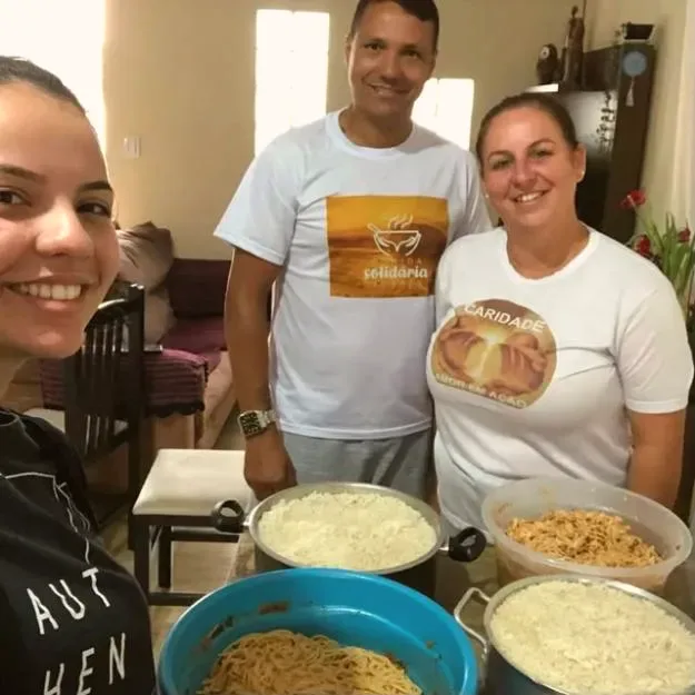Voluntários com camisetas Comida Solidária e Caridade ao lado de panelas de arroz, massa e frango